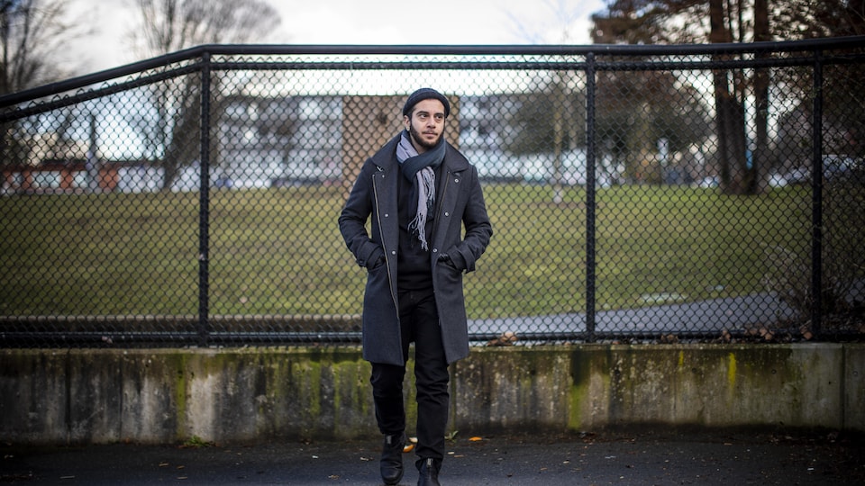 une personne debout dans la rue, devant un grillage, les mains dans les poches.