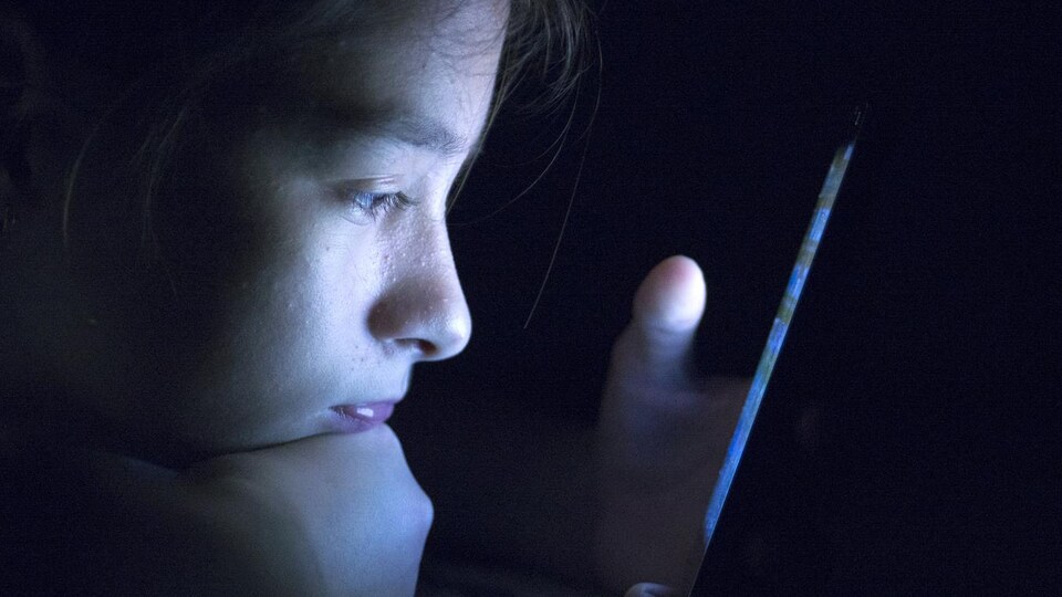 Un adolescent couché sur le ventre et appuyé sur son poing droit regarde l'écran de son téléphone dans le noir.