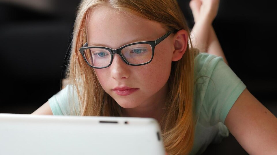 Une fillette avec des lunettes regarde l'écran d'une tablette. 