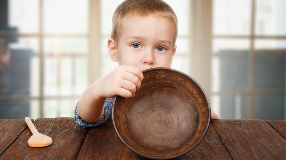 Un petit garçon, l'air sérieux, semble nous montrer un bol vide. Il est assis à une table en bois et une cuillère de bois est posée près de lui.