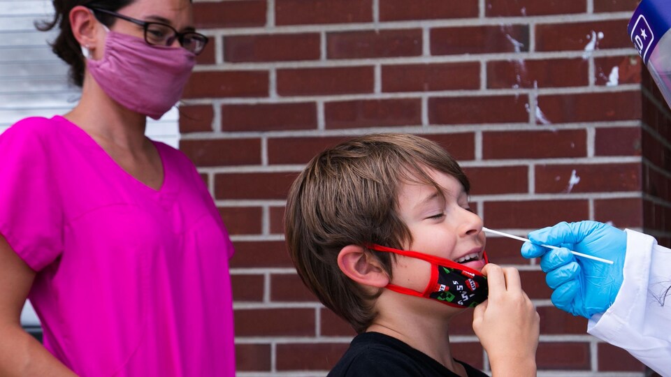 Un enfant tire sur son masque et fait la grimace alors que l'infirmière lui insère un écouvillon dans une narine.