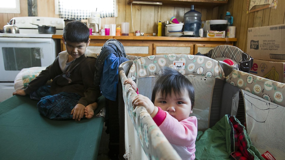 Deux enfants autochtones dans une maison. L'un est assis sur un lit de camp, l'autre est dans un lit pour bébé.