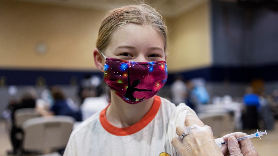 Une jeune fille qui porte un masque décoré d'étoiles scintillantes a un air heureux pendant qu'elle reçoit un vaccin.