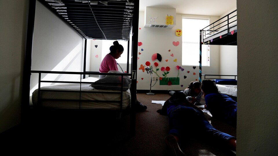 Dans une chambre avec deux lits superposés, une adolescente est assise sur l'un des matelas du bas alors que trois de ses camarades sont étendus par terre; il y a des décorations dessinées à la main sur le mur du fond.