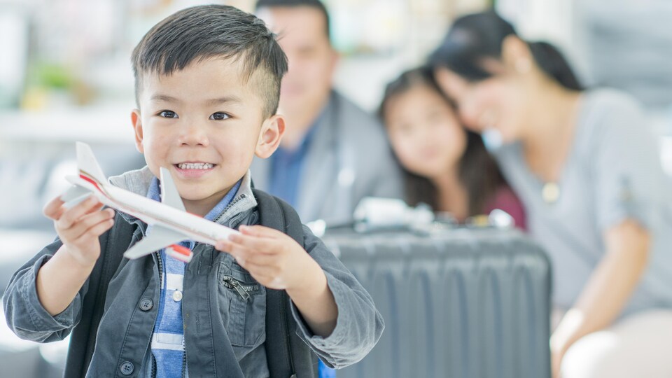 À l'avant-plan, un garçonnet asiatique à l'aéroport joue avec une réplique miniature d'avion avec sa famille derrière lui.
