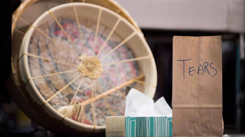 Un sac de papier brun sur lequel est écrit «larmes» repose à côté d'une boîte de papiers-mouchoirs et de deux tambours rituels autochtones en peau d'animal.