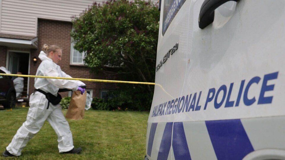 Une personne de l'équipe d'enquête marche sur la pelouse devant le domicile en transportant un sac de papier brun qui contient un élément de preuve.