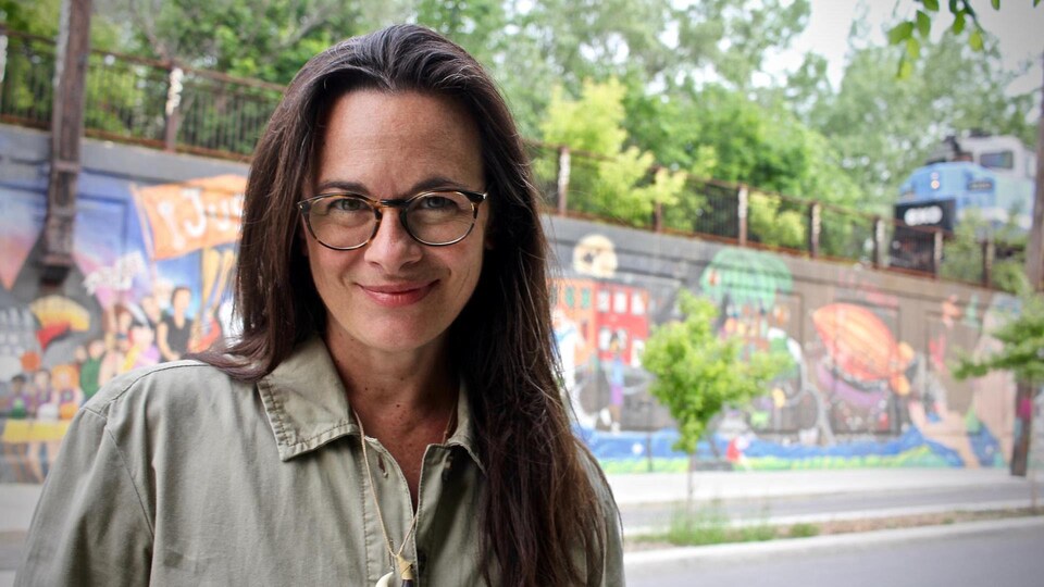 Une femme sourit et porte des lunettes. Derrière elle se trouve une murale colorée.