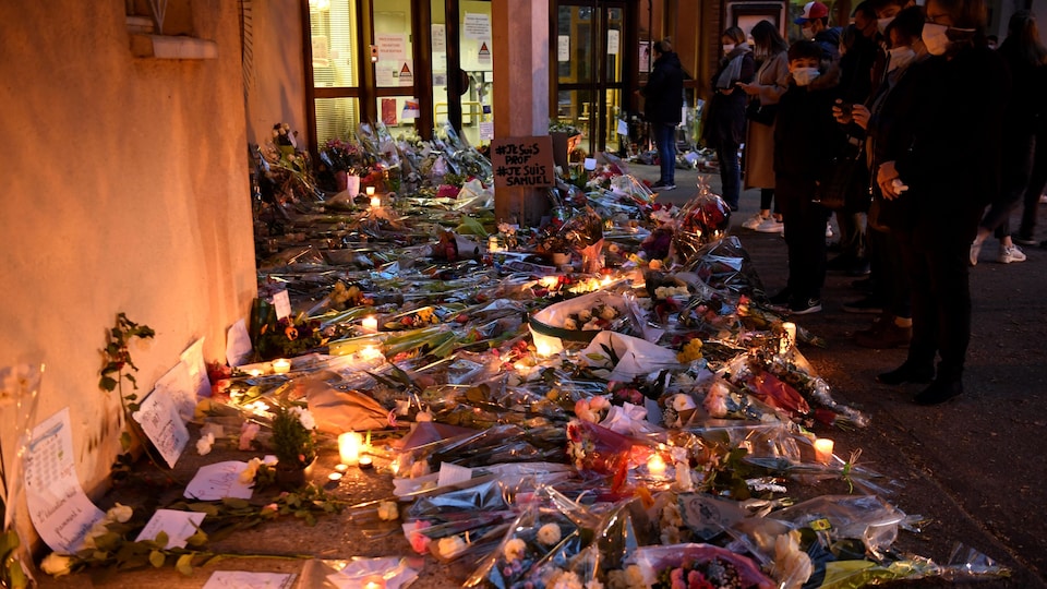 Des gens se tiennent debout devant des gerbes de fleurs, des chandelles allumées et des pancarte, dont l'une indique «#Je suis prof #Je suis Samuel».