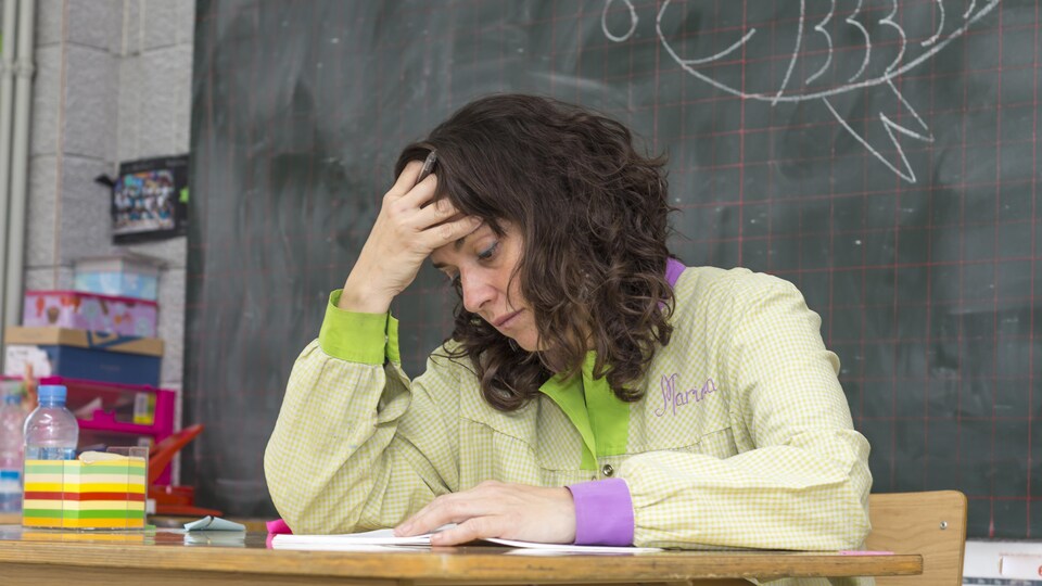 Une femme se tient la tête en regardant des documents, assise à un bureau dans une classe, avec le tableau derrière elle.