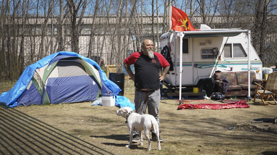 Guylain Levasseur se tient debout fièrement en compagnie de son chien, devant sa petite roulotte et sa tente.