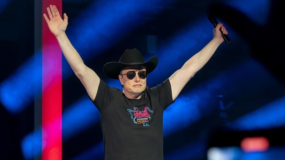 Elon Musk, lors d'un rassemblement, avec un chapeau de cowboy et des lunettes fumées, les mains en l'air.