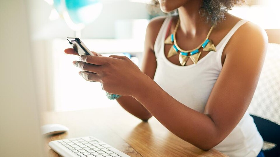 Une femme noire tient son portable, devant un ordinateur.