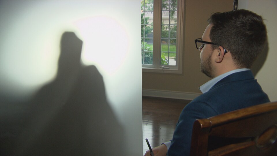 Le journaliste Jérôme Bergeron, assis sur une chaise, regarde devant lui un grand rideau blanc qui laisse entrevoir l'ombre d'une personne.