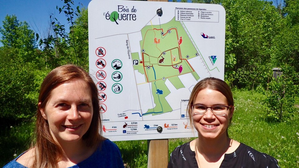 Virginie Dufour, responsable des dossiers de l'environnement au comité exécutif de la vile de Laval, et Louise Morin, consultante pour l'équipe Milieux naturels du Service de l'environnement de Laval. Elles posent devant une affiche qui indique les sentiers du Bois de l'Équerre