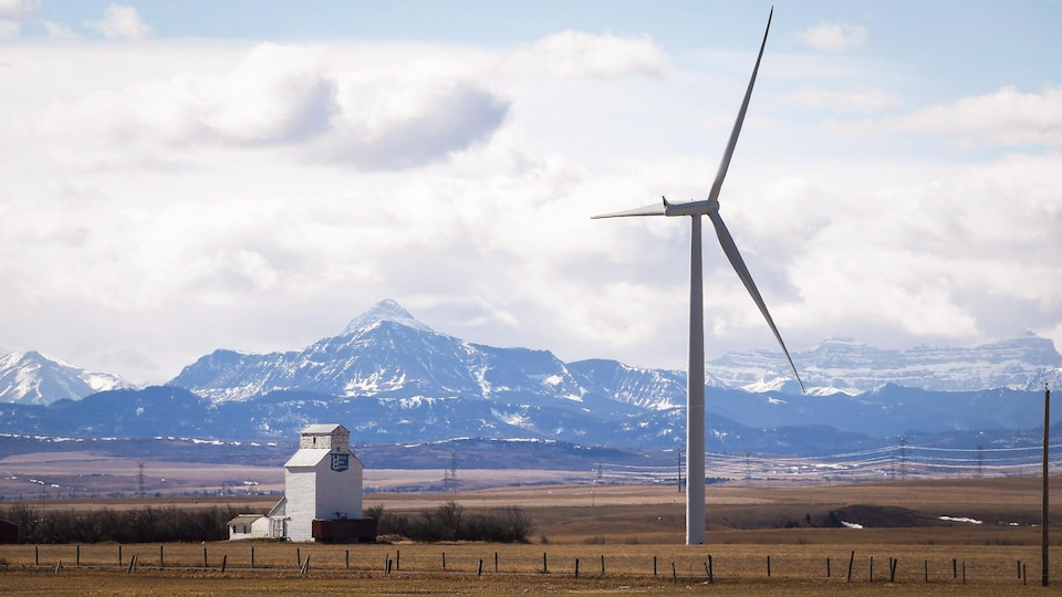 Paysage du sud de l'Alberta avec une éolienne en premier plan à côté d'un ascenseur à grain et les montagnes Rocheuses en arrière-plan