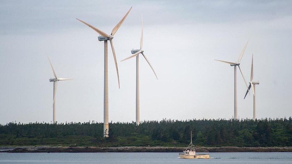 Cinq éoliennes surplombe une foret près d'un cours d'eau où un petit bateau de pêche se promène.  