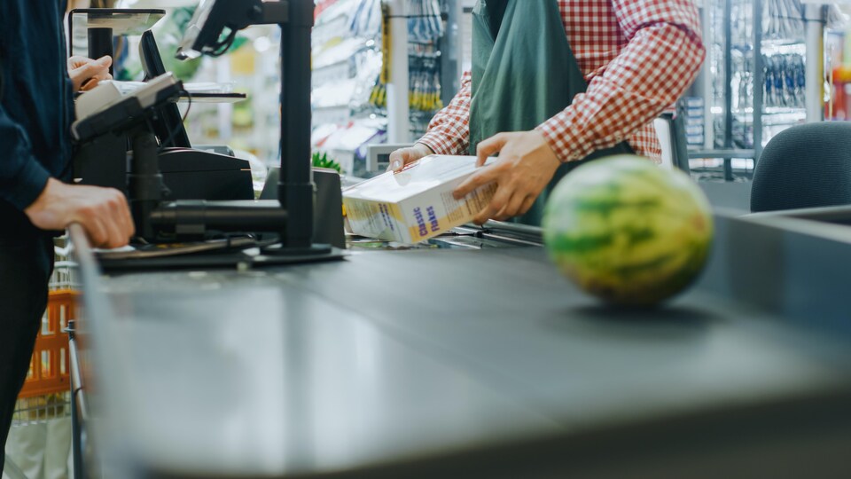 Un employé d'une épicerie passe les achats d'un client, dont un melon d'eau, devant un lecteur numérique.
