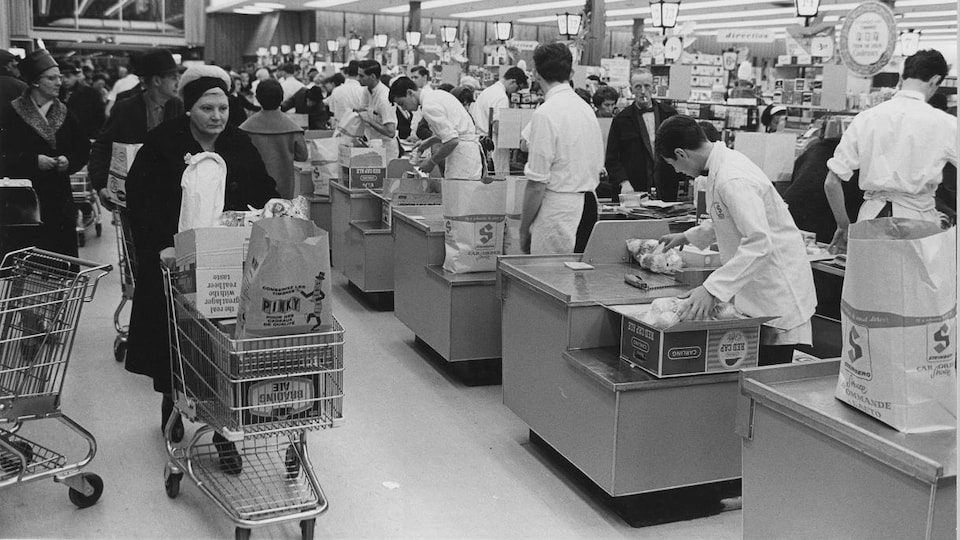 Une femme passe avec son panier d'épicerie rempli derrière la rangée de caisses d'un supermarché Steinberg.