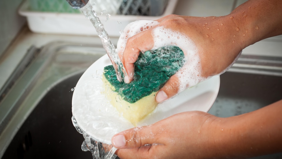 Une personne nettoie une assiette avec une éponge sous le robinet dans un lavabo de cuisine