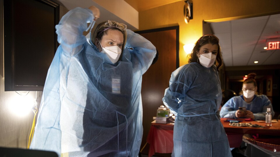 Deux infirmières debout, vêtues d'équipement de protection individuelle. L'une enfile une blouse d'hôpital. Une autre travailleuse de la santé vêtue de la même manière est assise plus loin derrière.