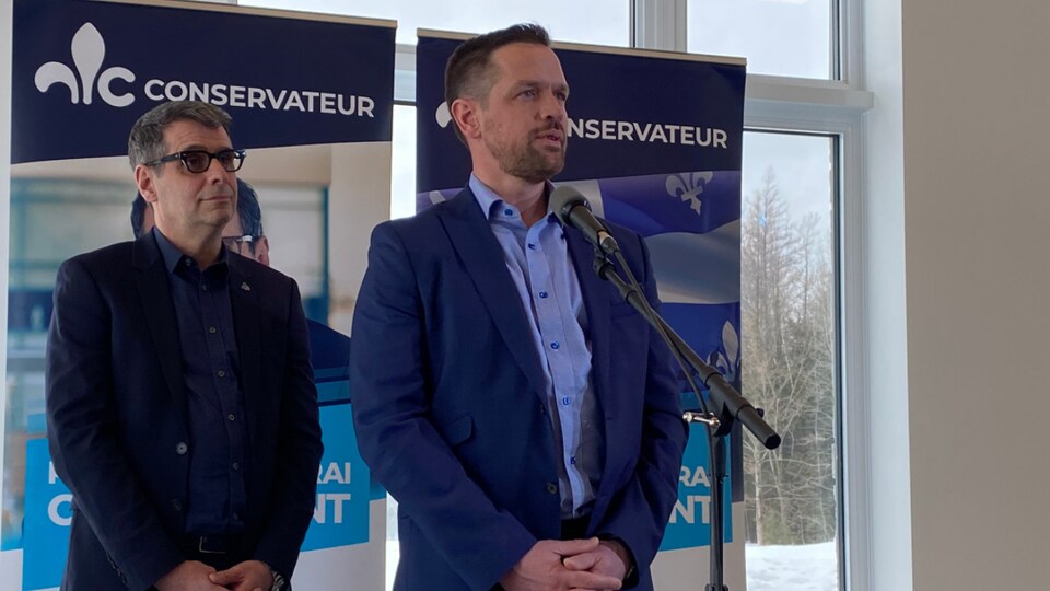 Deux hommes debout sur une scène avec des bannières du Parti conservateur du Québec derrière eux.