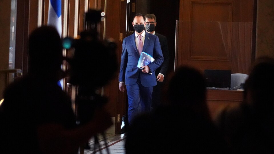 Deux hommes portant veston, cravate et masque de protection s'avancent dans une salle devant des caméras de télévision.
