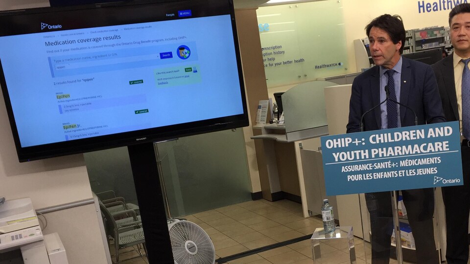 Le ministre Hoskins en conférence de presse dans une pharmacie, à côté d'un écran où est affiché le site web.