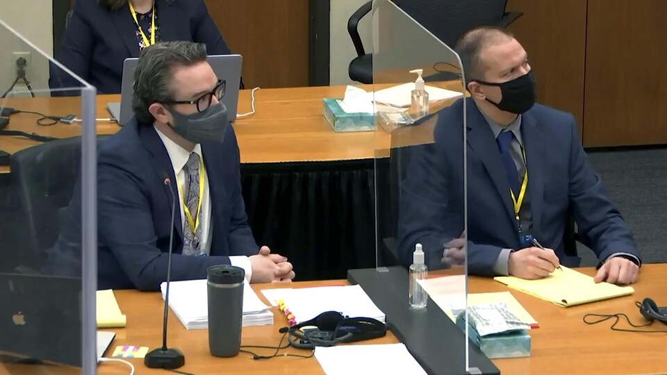 Deux hommes en complet, assis côte à côte et séparés par un plexiglas, au palais de justice.