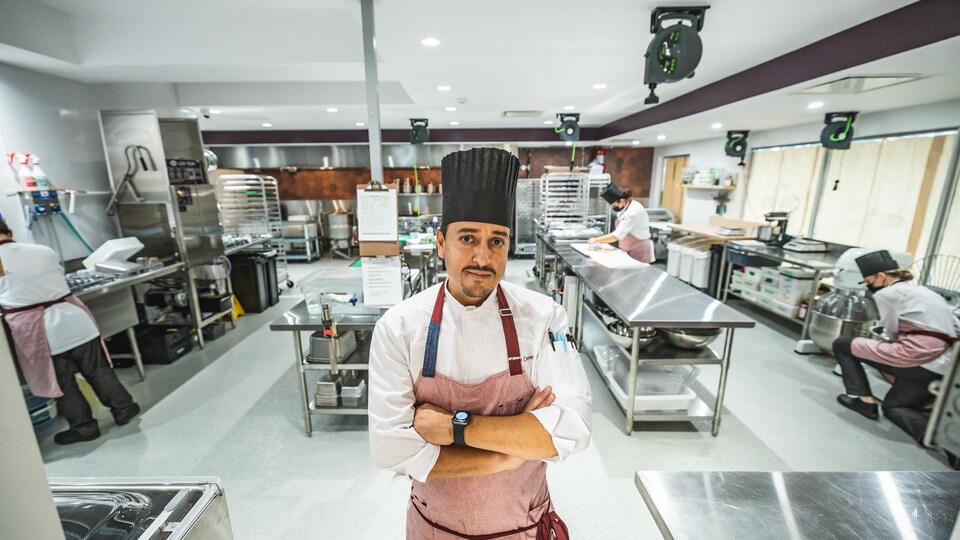 Un chef installé debout dans une grande cuisine d'un restaurant, le regard fier, avec des collègues qui travaillent derrière lui.