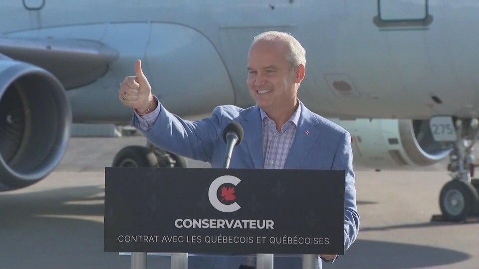 Le chef conservateur Erin O'Toole est derrière un lutrin et lève le pouce droit, avec un avion à l'arrière-plan.