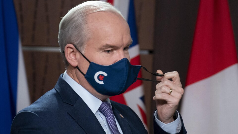 Devant les drapeaux du Canada et des provinces, Erin O'Toole ôte son masque à l'effigie du Parti conservateur du Canada.
