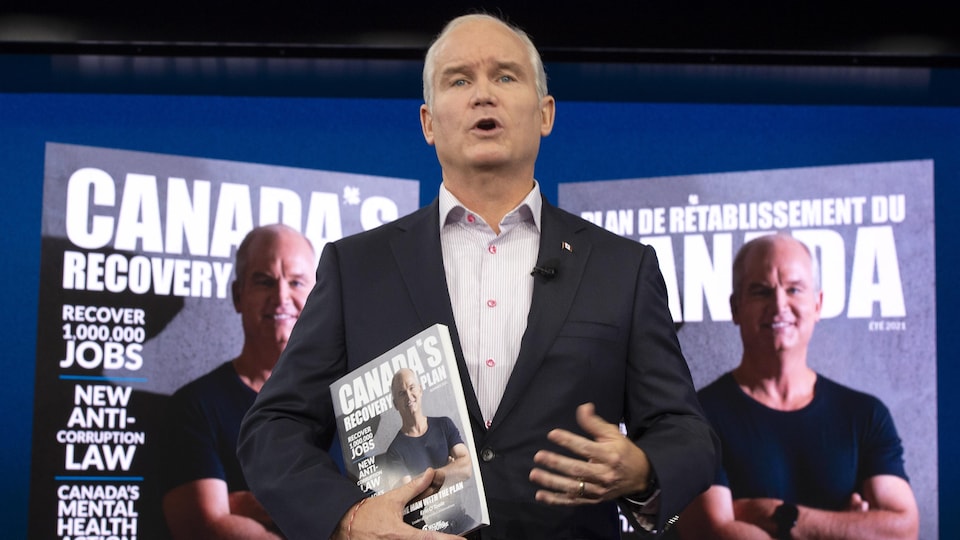 Erin O'Toole tient le programme conservateur dans sa main, tandis que la page couverture le présentant les bras croisés en t-shirt est projetée en anglais et en français derrière lui.