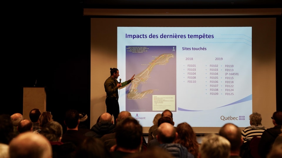 Un homme devant une projection sur la scène avec dans la salle des gens qui écoutent.