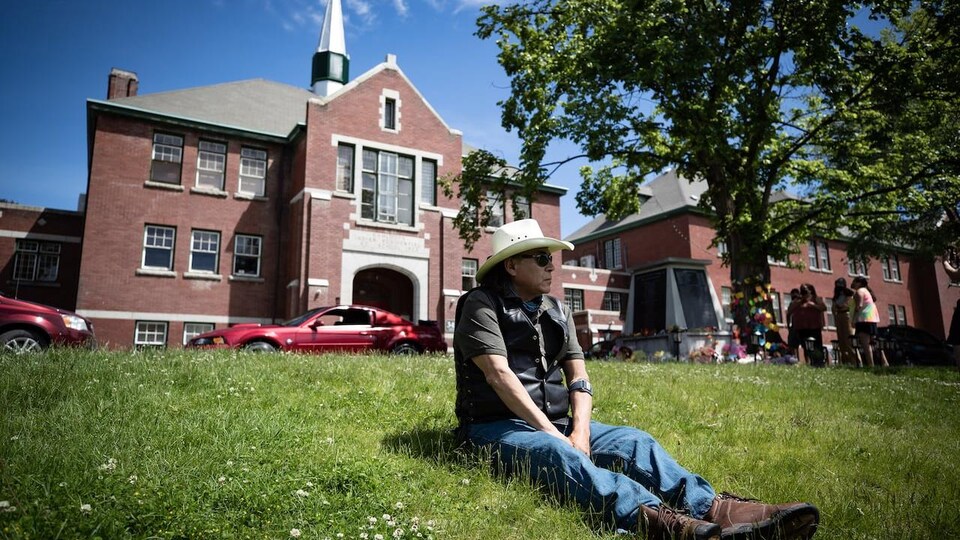 Un homme portant un chapeau de cowboy est assis sur l'herbe devant un bâtiment.