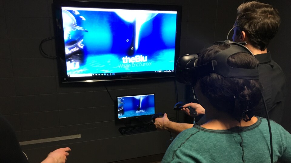 Une femme a un casque de réalité virtuelle sur les yeux, pendant qu'un homme contrôle un ordinateur avec des manettes.