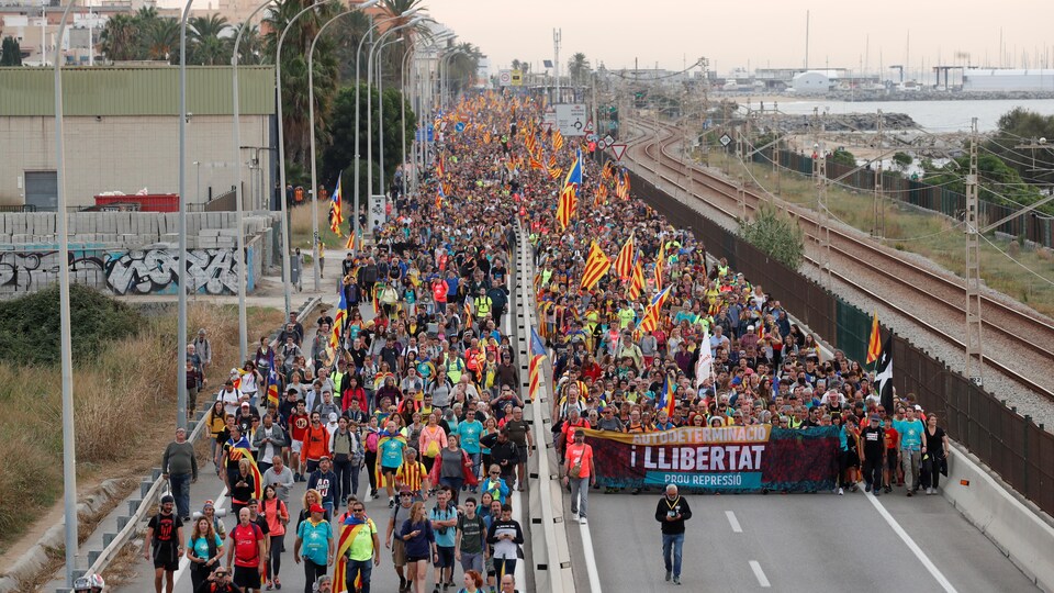Un long cortège de manifestants s'étend au loin. Des drapeaux catalans jaune, rouge et bleu flottent au vent.