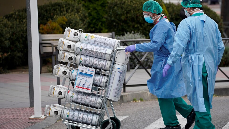 Deux travailleurs de la santé, portant des masques de protection, transportent des bouteilles d'oxygène sur un chariot.