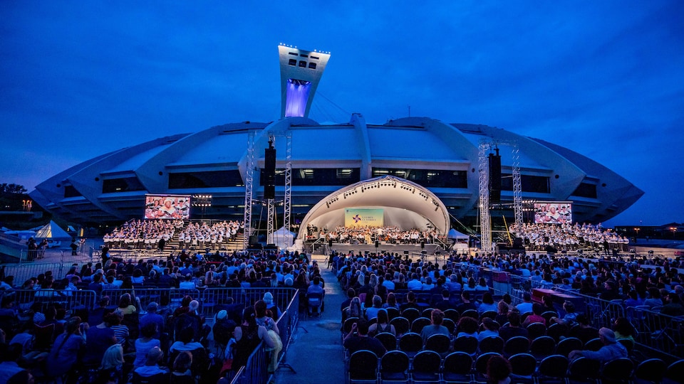 L'Orchestre symphonique de Montréal joue dans un amphithéâtre érigé devant le stade olympique. Des milliers de spectateurs et spectatrices assistent au concert.