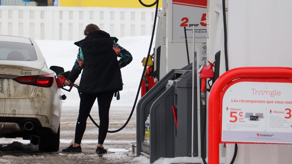 Une femme de dos dans une station-essence en train de faire le plein de sa voiture. Il y a de la neige au sol.