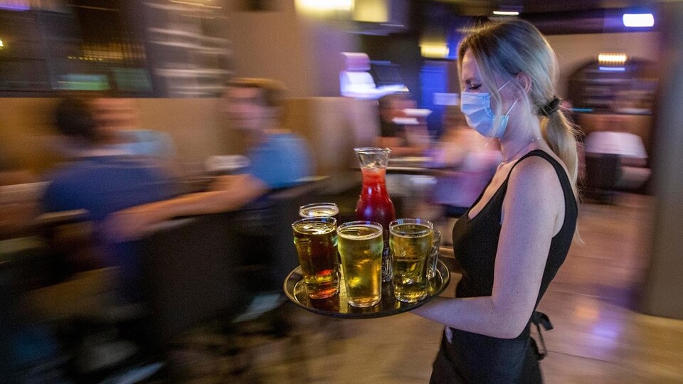 Une serveuse avec un masque sur le visage porte un plateau recouvert de boissons dans une salle de restaurant.