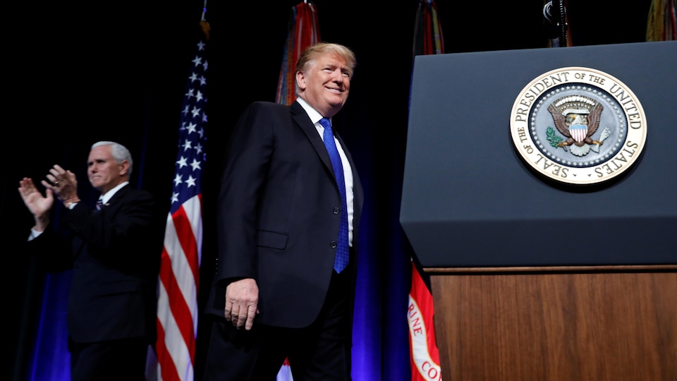 Le président américain Donald Trump, souriant à côté d'un podium, alors qu'on voit Mike Pence qui applaudit à l'arrière.