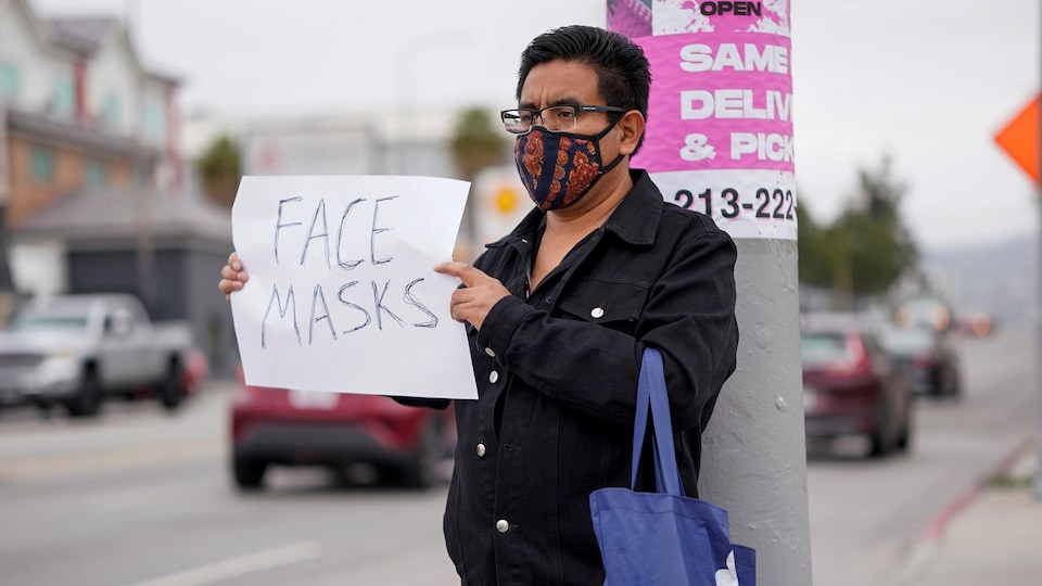 Un homme dont le visage est couvert d'un masque en tissu tient un écriteau sur lequel est écrit « Masques pour le visage».