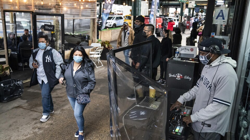 Un DJ qui porte un masque joue dans un abri protégé par du plexiglas dans la rue. Des dizaines de personnes se promènent dans la rue.