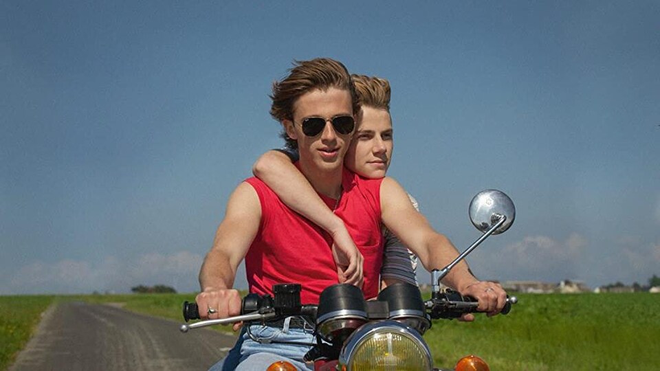 Deux jeunes sont sur une moto, cheveux au vent, sur une route de campagne.