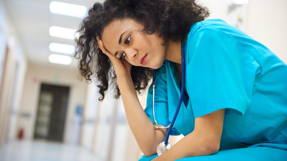 Une infirmière assise se tient la tête dans un corridor d'hôpital.