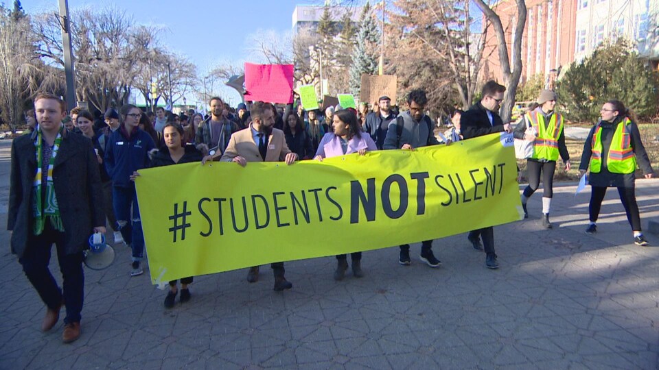 Des étudiants marchent en rangs. Le premier rang tient une large banderole. Il y est écrit en anglais les étudiants ne seront pas silencieux.