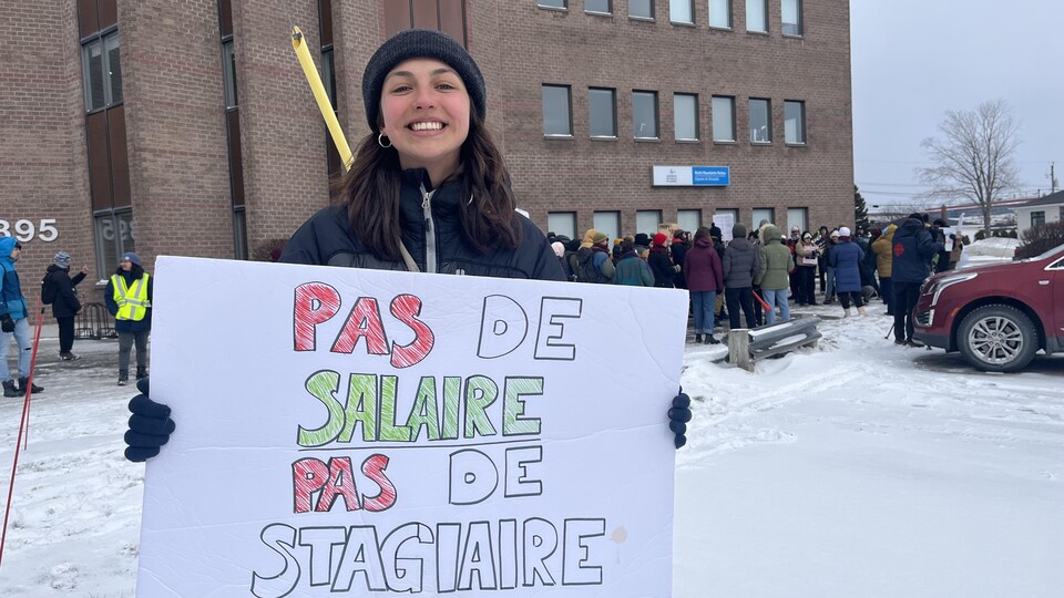 Une étudiante sourit en tenant une pancarte sur laquelle on peut lire : « Pas de salaire, pas de stagiaire ». 