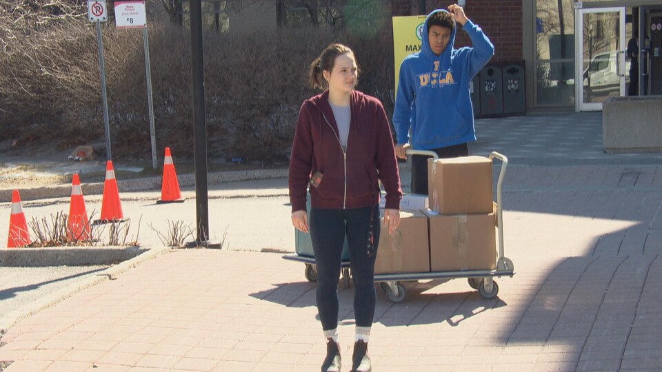 Deux étudiants attendent devant un bâtiment avec des caisses posées sur un charriot