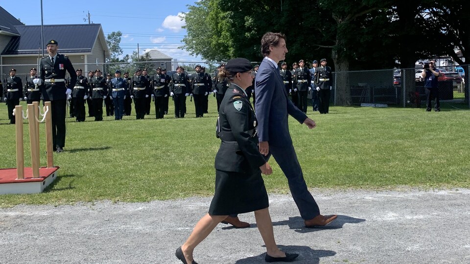 Justin Trudeau fait son entrée. Il marche sur un chemin de gravier dans le parc, accompagné d'une militaire en uniforme.